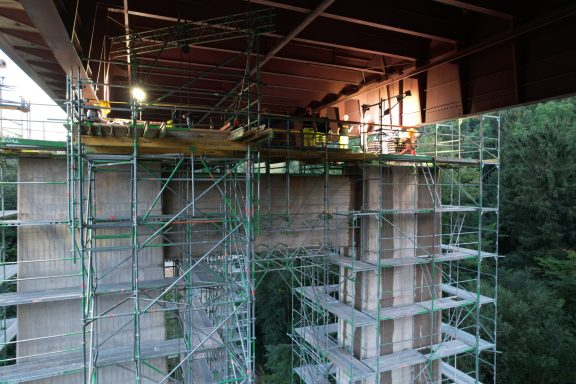 Bauarbeiten an einer Brücke mit Gerüst und Betonpfeilern in einer grünen Umgebung.