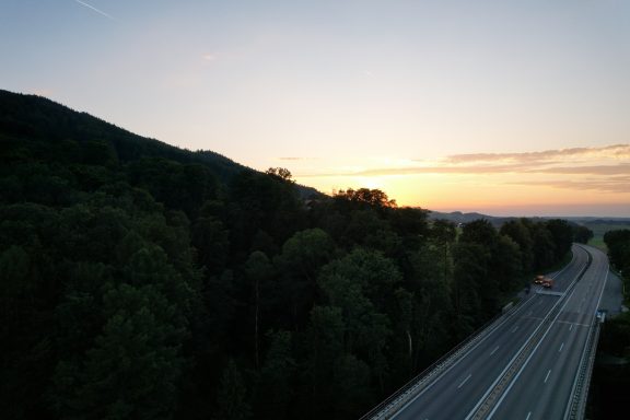 Sonnenuntergang über einer Landstraße, umgeben von bewaldeten Hügeln.