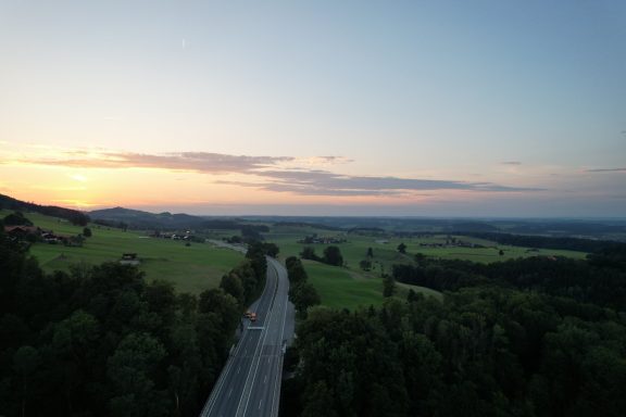 Blick auf eine kurvenreiche Straße durch grüne Felder bei Sonnenuntergang.